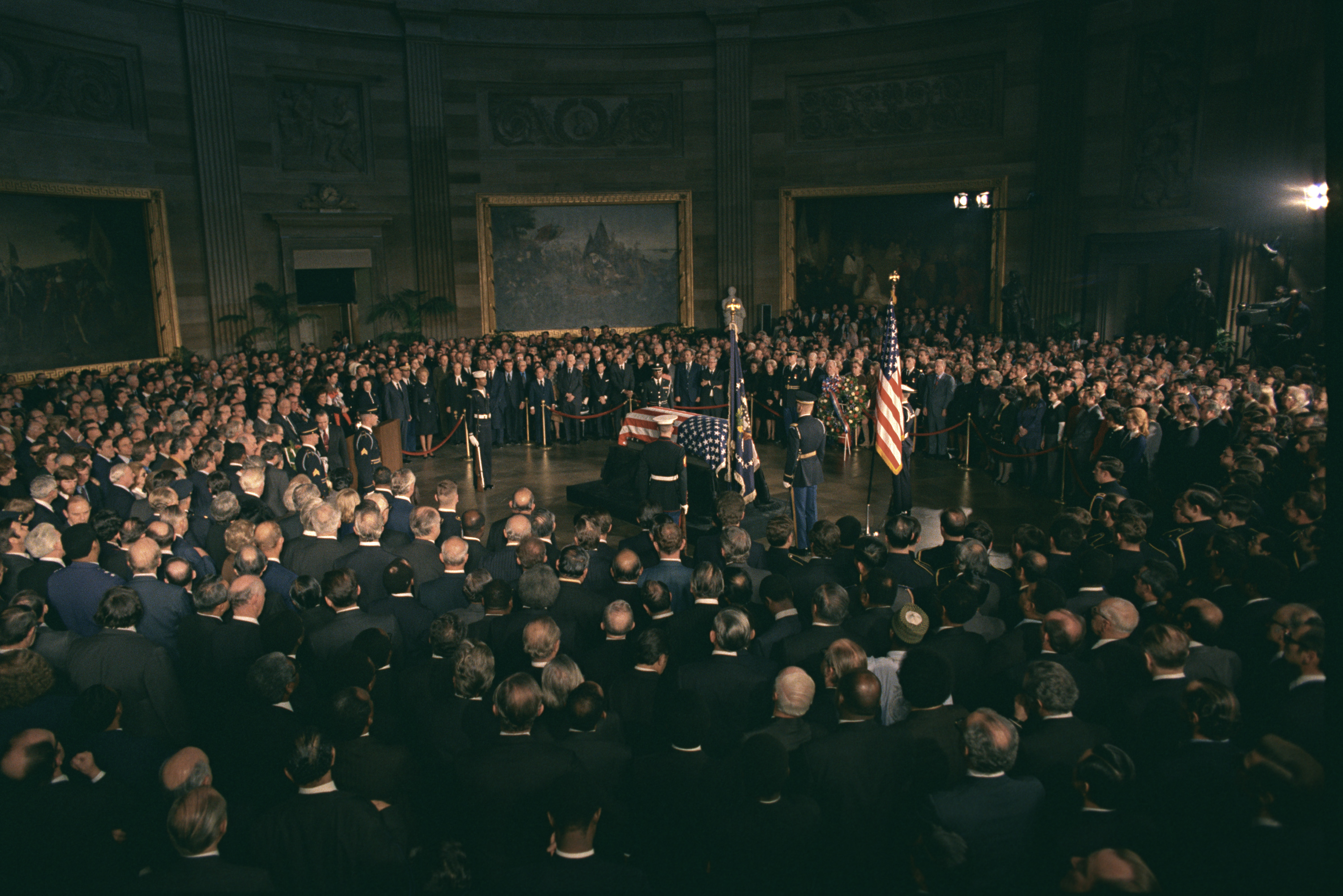President Richard M. Nixon, members of Congress, and others paying respects to President Lyndon B. Johnson as he lies in state. The casket, draped in an American flag sits in the center of a room, a crowd encircling it.